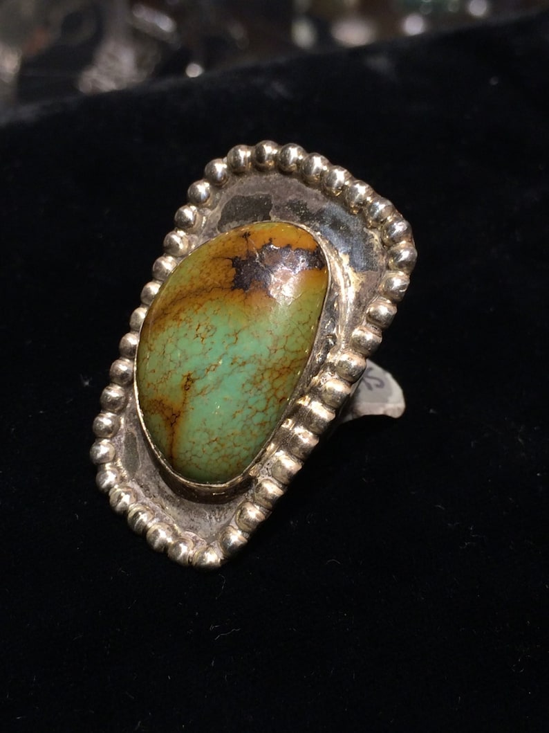 Pu&ograve; includere: Un anello d'argento con una pietra turchese verde e marrone incastonata in una lunetta a forma di conchiglia. L'anello &egrave; in argento sterling e ha una finitura testurizzata.