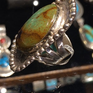 Pu&ograve; includere: Un anello d'argento con una grande pietra ovale di turchese incastonata in una montatura a castone. L'anello ha un bordo perlato e una fascia decorativa.