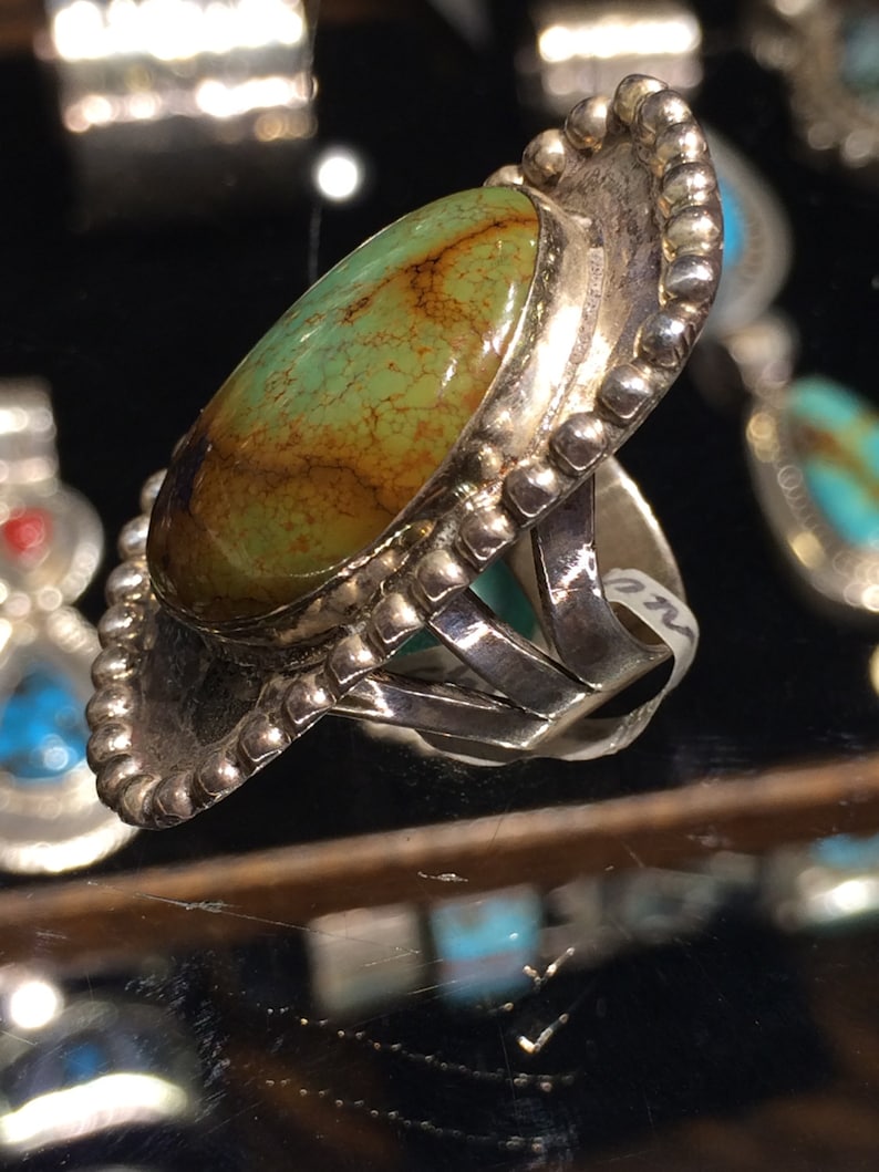 Pu&ograve; includere: Un anello d'argento con una grande pietra ovale di turchese incastonata in una montatura a castone. L'anello ha un bordo perlato e una fascia decorativa.