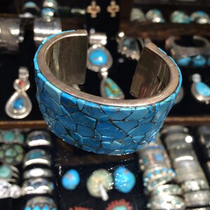 May include: A silver cuff bracelet with a mosaic of turquoise stones. The bracelet is made of a silver metal and has a smooth, polished finish. The turquoise stones are set in a geometric pattern and are a vibrant blue color.