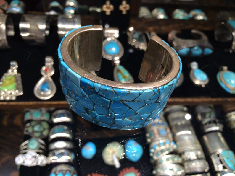 May include: A silver cuff bracelet with a mosaic of turquoise stones. The bracelet is made of a silver metal and has a smooth, polished finish. The turquoise stones are set in a geometric pattern and are a vibrant blue color.