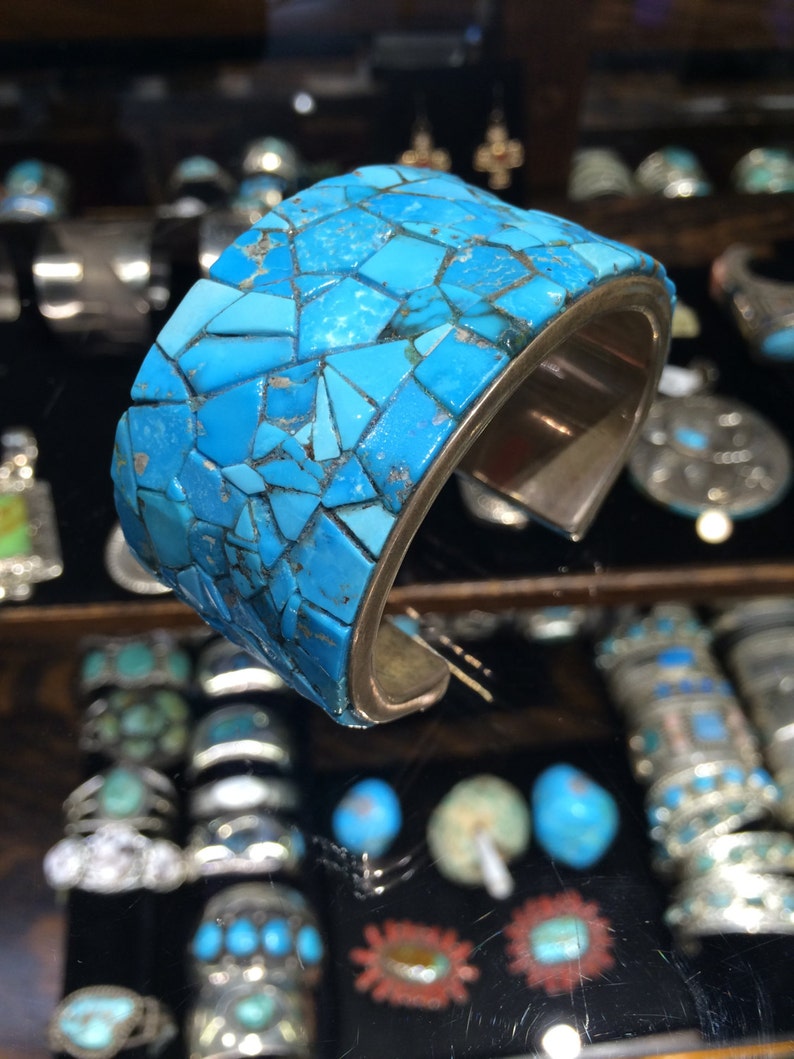May include: A turquoise and silver cuff bracelet with a mosaic design. The bracelet is made of many small turquoise stones that are set in silver. The bracelet is a beautiful example of Native American jewelry.