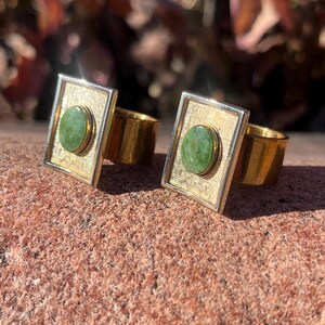 May include: Two gold rings with square frames and green oval stones. The rings are on a brown textured surface.