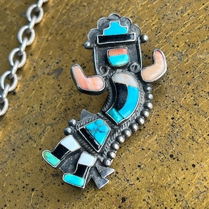 May include: A silver pendant in the shape of a stylized figure, inlaid with turquoise, black, white, and coral-colored stones. The pendant has a detailed design and is attached to a silver chain. The figure has outstretched arms and detailed feet.