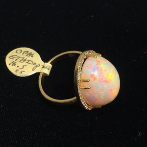 Op de afbeelding: Een gouden ring met een grote ovale opaal-edelsteen gezet in een bezel-setting met een halo van kleine diamanten. De opaal is melkachtig wit met schittering in roze, oranje en groen.