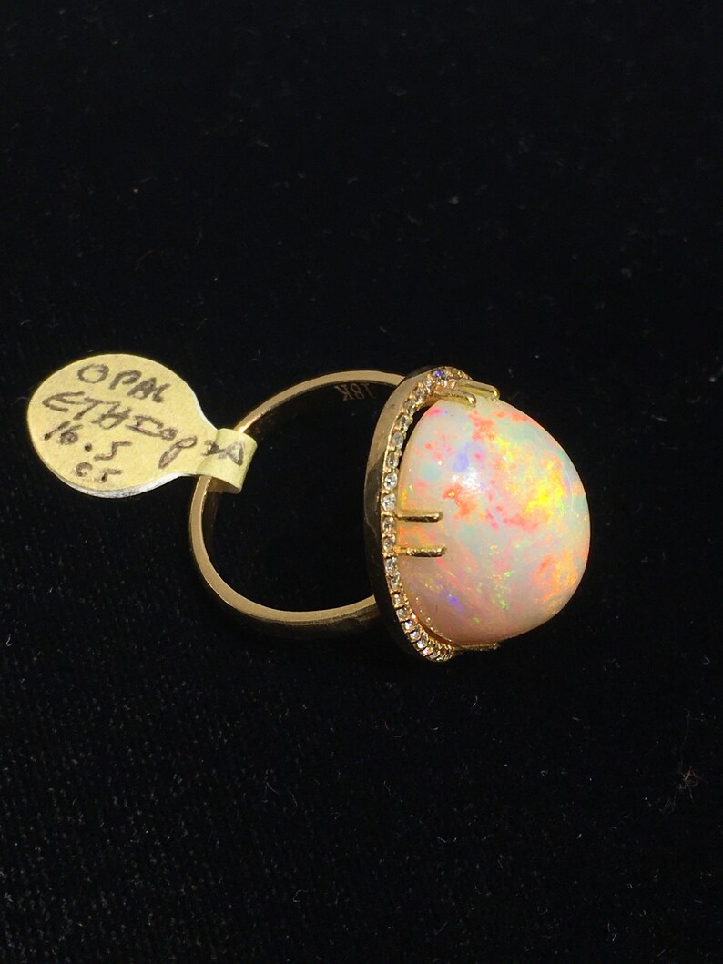 Op de afbeelding: Een gouden ring met een grote ovale opaal-edelsteen gezet in een bezel-setting met een halo van kleine diamanten. De opaal is melkachtig wit met schittering in roze, oranje en groen.
