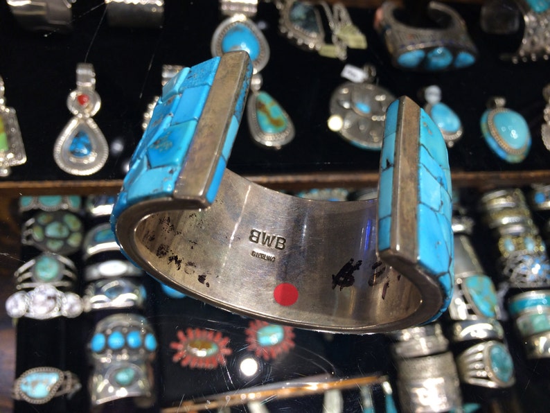 May include: A silver cuff bracelet with turquoise inlay. The bracelet is decorated with a geometric pattern and has a silver band with the inscription "BWB STERLING".