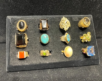 May include: A display of 12 gold rings with various gemstones, including black, brown, turquoise, blue, green, and clear stones. The rings are arranged in a grid pattern on a black velvet background.