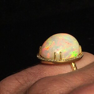 Op de afbeelding: Een gouden ring met een grote, witte opaal-edelsteen die in een bezel-setting is gezet. De opaal heeft een regenboog aan kleuren, waaronder rood, oranje, geel, groen, blauw en paars. De ring is versierd met kleine diamanten rond de bezel.