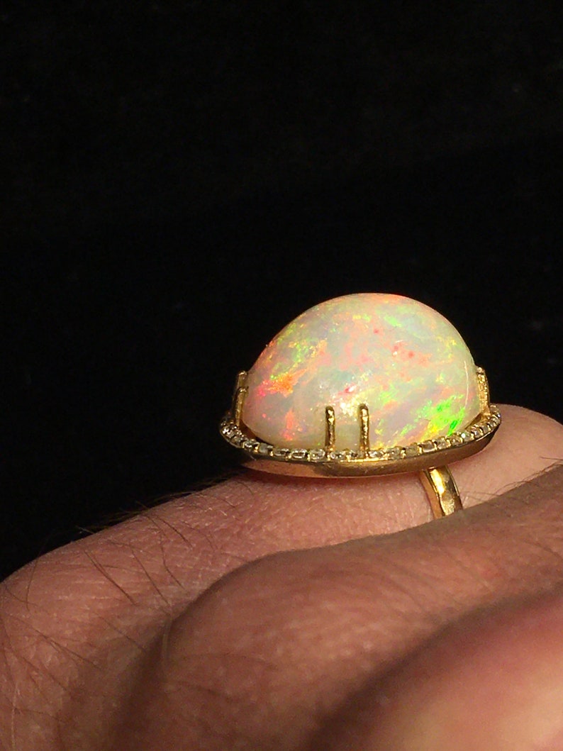 Op de afbeelding: Een gouden ring met een grote, witte opaal-edelsteen die in een bezel-setting is gezet. De opaal heeft een regenboog aan kleuren, waaronder rood, oranje, geel, groen, blauw en paars. De ring is versierd met kleine diamanten rond de bezel.