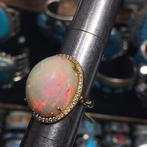 Op de afbeelding: Een gouden ring met een grote ovale opaal-edelsteen omringd door een halo van kleine diamanten. De opaal heeft een witte basis met roze en rode flitsen.