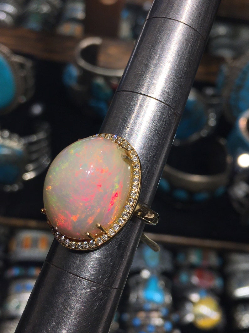 Op de afbeelding: Een gouden ring met een grote ovale opaal-edelsteen omringd door een halo van kleine diamanten. De opaal heeft een witte basis met roze en rode flitsen.