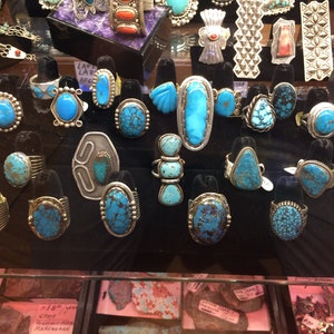 May include: A display of turquoise rings set in silver. The rings feature various styles and sizes of turquoise stones, including round, oval, and rectangular shapes. The stones are a vibrant blue-green color and have a variety of patterns and textures.