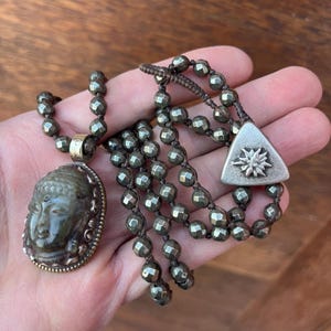 May include: A necklace featuring a Buddha pendant and a separate strand of faceted beads. The Buddha pendant is oval-shaped with a detailed face and a gold-colored frame. The beads are a dark, metallic gray color.