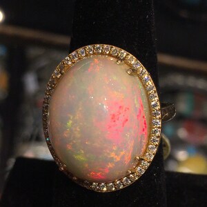 Op de afbeelding: Een ring met een grote ovale opaal-edelsteen omringd door een halo van kleine diamanten. De opaal is melkachtig wit met schittering in roze, groen en geel.