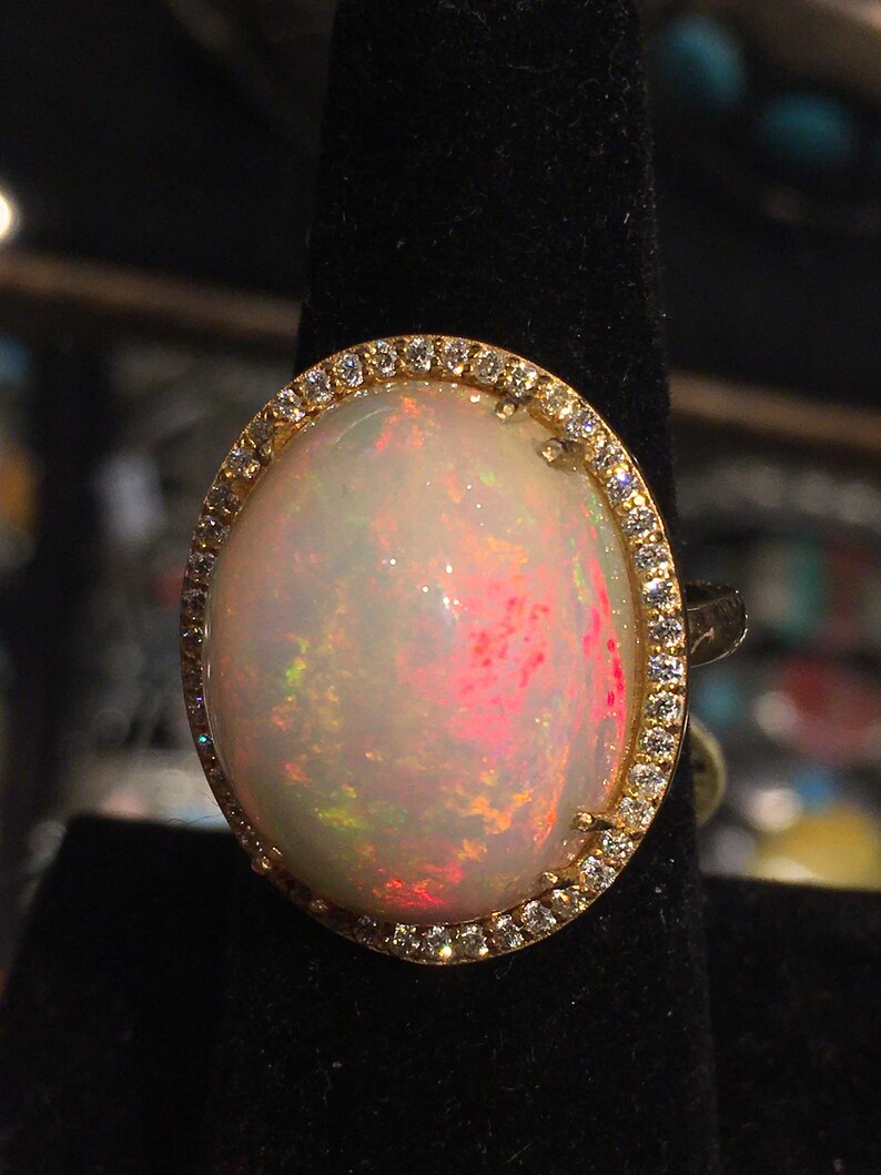 Op de afbeelding: Een ring met een grote ovale opaal-edelsteen omringd door een halo van kleine diamanten. De opaal is melkachtig wit met schittering in roze, groen en geel.