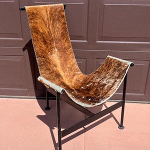 May include: A mid-century modern chair with a cowhide seat. The seat is brown and white with a curved design. The frame is black metal with four legs. The chair is designed for comfort and style.