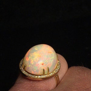 Op de afbeelding: Een gouden ring met een grote, ovale opaal-edelsteen gezet in een halo van kleinere diamanten. De opaal heeft een witte basis met flitsen van oranje, geel en groen.
