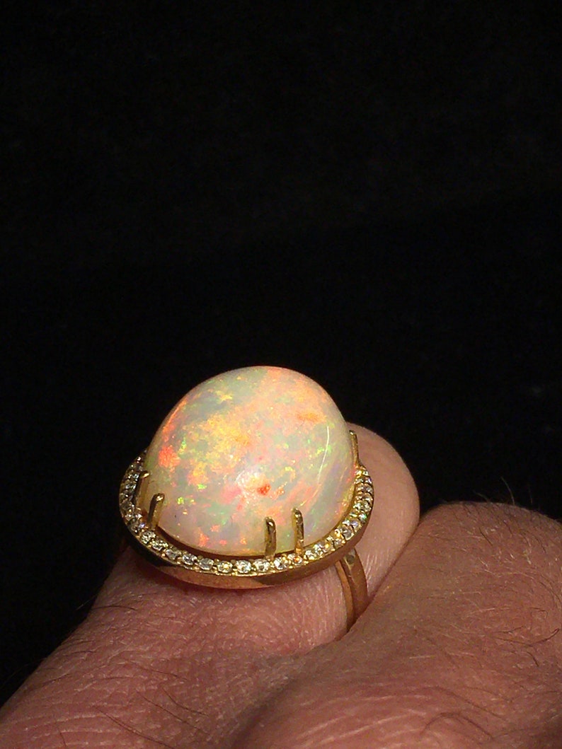 Op de afbeelding: Een gouden ring met een grote, ovale opaal-edelsteen gezet in een halo van kleinere diamanten. De opaal heeft een witte basis met flitsen van oranje, geel en groen.