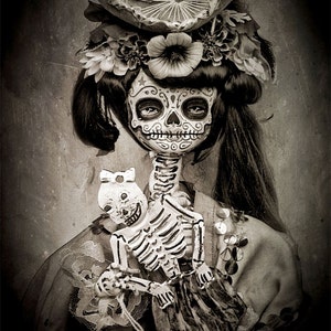 May include: A woman in a black and white costume with a sugar skull face paint, wearing a sombrero and holding a skeleton doll. The background is a textured, faded black and white image.
