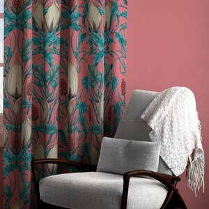 Op de afbeelding: Een paar roze gordijnen met een bloemenpatroon met witte bloemen en groene bladeren. De gordijnen hangen voor een raam met witte lijst en uitzicht op een bewolkte lucht. Een grijze fauteuil met een witte deken staat op de voorgrond.