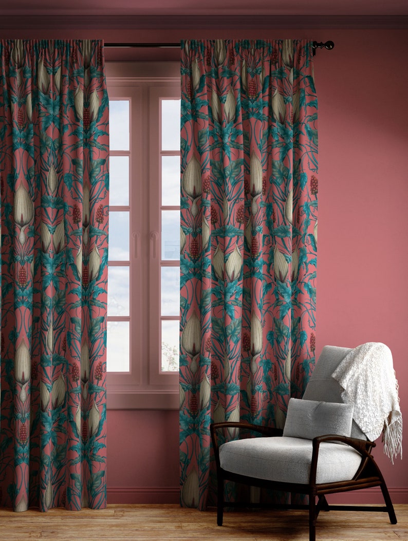 Op de afbeelding: Een paar roze gordijnen met een bloemenpatroon met witte bloemen en groene bladeren. De gordijnen hangen voor een raam met witte lijst en uitzicht op een bewolkte lucht. Een grijze fauteuil met een witte deken staat op de voorgrond.