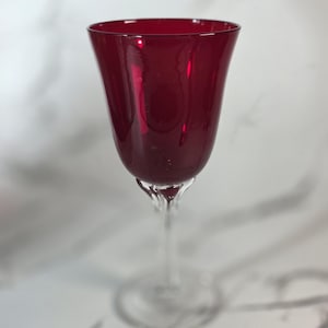 May include: A ruby red wine glass with a tulip-shaped bowl and a slender stem. The glass is made of transparent material, allowing light to pass through. The glass is set against a white marble background.