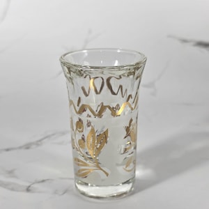 May include: Clear glass shot glass with gold floral and geometric designs. The shot glass has a textured surface and a slightly flared rim. The gold design includes floral and geometric patterns. The glass is approximately 3 inches tall.