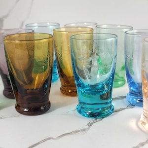 May include: A set of ten colorful glass shot glasses with various etched designs. The glasses are arranged in a row on a white surface.