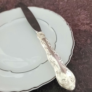 May include: A silver butter knife with an ornate handle on a white plate with a black rim.