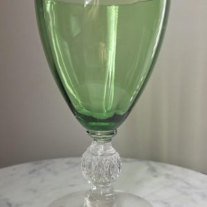 May include: A green-tinted glass goblet with a clear, textured stem and a wide, round base. The bowl of the glass is a vibrant green, and the stem features a decorative, textured design. The goblet is designed for beverages.