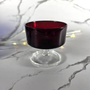 May include: A burgundy-colored glass dessert cup with a clear, circular base and stem. The cup has a wide, rounded bowl. The glass is smooth and reflective, with a slight transparency. The cup is likely used for serving desserts or small portions.