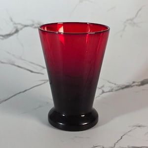 May include: A red glass vase with a flared top and a rounded base. The color transitions from a deep red at the top to a dark, almost black, shade at the base. The vase is empty and sits against a white marble background.