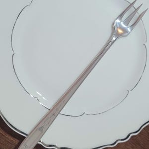 May include: A silver cake fork with a decorative handle on a white plate with a scalloped edge.