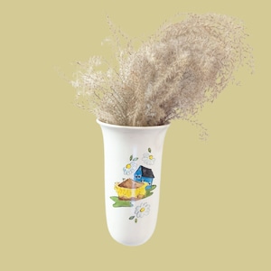 Puede incluir: Jarrón de cerámica blanco con un diseño pintado en azul y amarillo de una casa y un barco. El jarrón está lleno de hierba de la pampa seca.