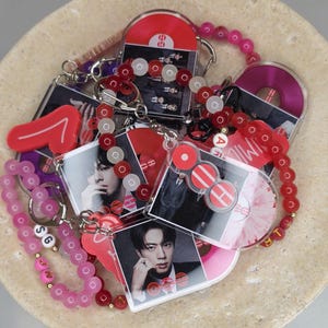 May include: A collection of acrylic keychains featuring photographic portraits of a person and vinyl record graphics. The keychains are attached to beaded chains in shades of red and pink, with some beads displaying the letters OOH, SG, and V.