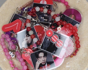 May include: A collection of acrylic keychains featuring photographic portraits of a person and vinyl record graphics. The keychains are attached to beaded chains in shades of red, pink, and white, with some beads displaying letters like SG, J, and V.