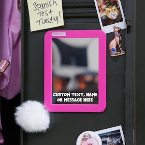 May include: A pink magnetic mirror with a white frame. The mirror is attached to a black metal surface. The text "CUSTOM TEXT, NAME OR MESSAGE HERE" is printed on the mirror.