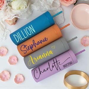 Puede incluir: Cuatro vasos personalizados en azul, naranja, gris y lavanda, cada uno con un nombre en un color contrastante. El vaso azul dice "Dillion", el naranja dice "Stephanie", el gris dice "Arianna" y el lavanda dice "Charlotte". Cada uno tiene una pajita a juego.