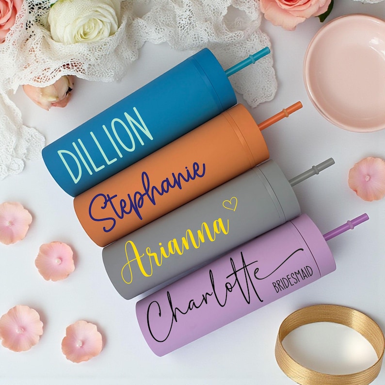 Puede incluir: Cuatro vasos personalizados en azul, naranja, gris y lavanda, cada uno con un nombre en un color contrastante. El vaso azul dice "Dillion", el naranja dice "Stephanie", el gris dice "Arianna" y el lavanda dice "Charlotte". Cada uno tiene una pajita a juego.