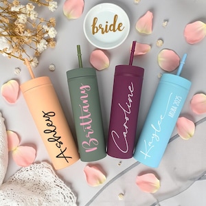 Four personalized acrylic skinny tumblers with straws, each in a different color (peach, sage green, plum, and light blue), displayed on a soft bridal-themed background with rose petals, lace, and a dish labeled "bride." Tumblers feature names in script fonts, ideal for bridesmaid or bachelorette gifts.