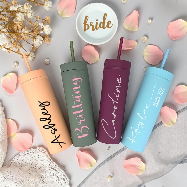 Four personalized acrylic skinny tumblers with straws, each in a different color (peach, sage green, plum, and light blue), displayed on a soft bridal-themed background with rose petals, lace, and a dish labeled "bride." Tumblers feature names in script fonts, ideal for bridesmaid or bachelorette gifts.