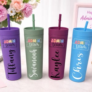 May include: Four colorful tumblers with straws, each personalized with a name and the text "ADMIN team." The tumblers are purple, green, burgundy, and blue. A floral arrangement and a "Happy Admin Day!" sign are in the background.