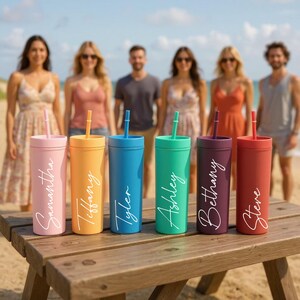 Puede incluir: Seis vasos de colores con pajitas est&aacute;n colocados sobre una mesa de madera en la playa. Cada vaso es de un color diferente: rosa, naranja, azul, verde, morado y rojo. Cada vaso tiene un nombre en escritura blanca: Samantha, Tiffany, Tyler, Ashley, Bethany y Steve.