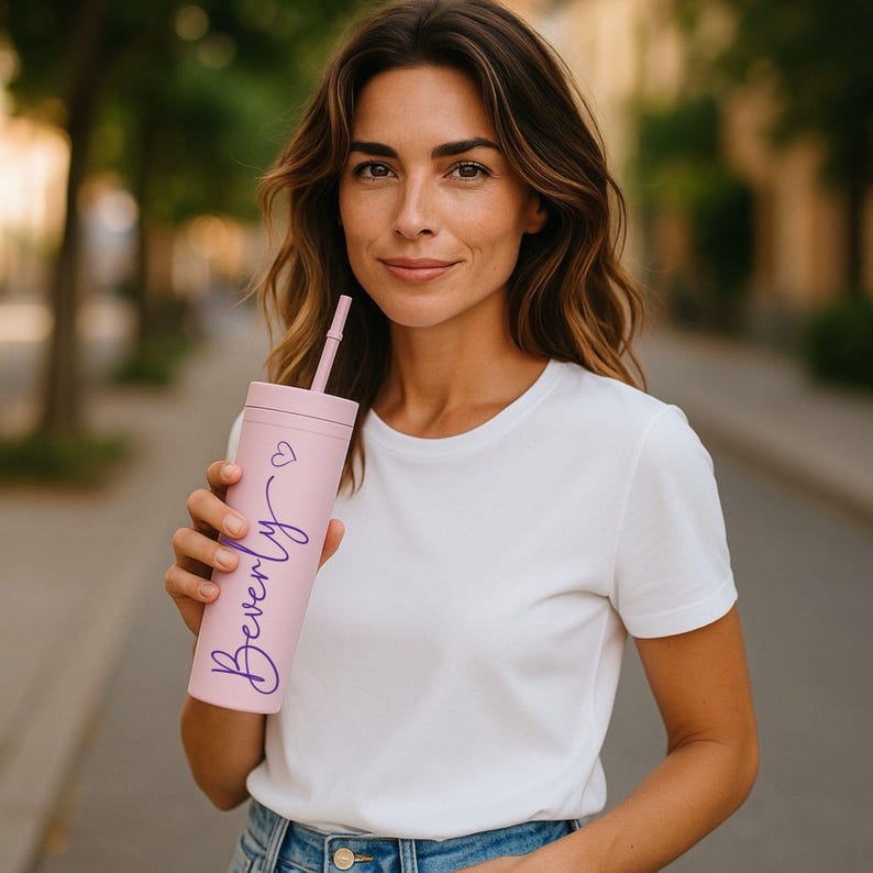 Puede incluir: Un vaso de color rosa claro con una pajita, con el nombre "Beverly" en una fuente p&uacute;rpura, junto con un peque&ntilde;o s&iacute;mbolo de coraz&oacute;n. El vaso es sostenido por una persona que lleva una camiseta blanca.
