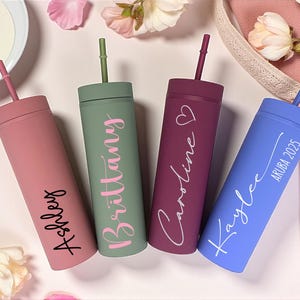 Puede incluir: Cuatro vasos delgados personalizados en rosa, verde, burdeos y azul, cada uno con una pajita a juego. Los vasos est&aacute;n inscritos con nombres y una fecha. El fondo incluye flores y una tela rosa.