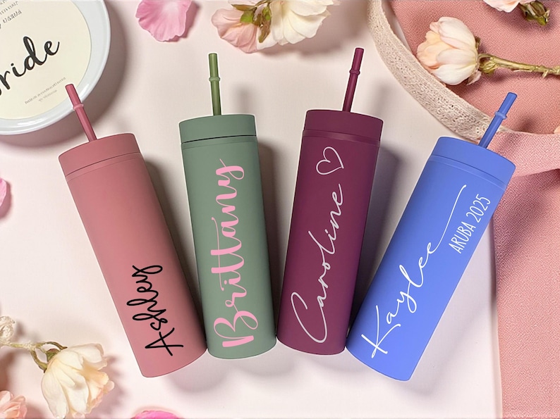 Puede incluir: Cuatro vasos delgados personalizados en rosa, verde, burdeos y azul, cada uno con una pajita a juego. Los vasos est&aacute;n inscritos con nombres y una fecha. El fondo incluye flores y una tela rosa.