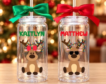May include: Two clear glass tumblers with lids and straws. The left tumbler features a green bow, the name KAITLYN in green, and a reindeer with a red bow. The right tumbler has a red bow, the name MATTHEW in red, and a reindeer with sunglasses.