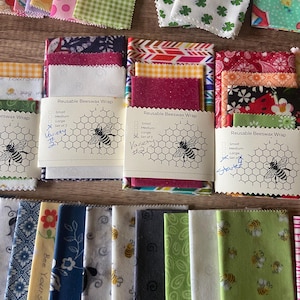 May include: A collection of reusable beeswax food wraps in various colors and patterns. The wraps are labeled with "Reusable Beeswax Wrap" and have a honeycomb design with a bee illustration. The labels also list different sizes: small, medium, large, and set of 3. Some of the wraps have handwritten notes on them, such as "Variety" and "Started".