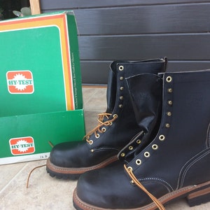 May include: A pair of black leather work boots with brown laces and gold eyelets. The boots are sitting on a tiled floor next to a green and orange box with the text "Hy-Test" printed on it.
